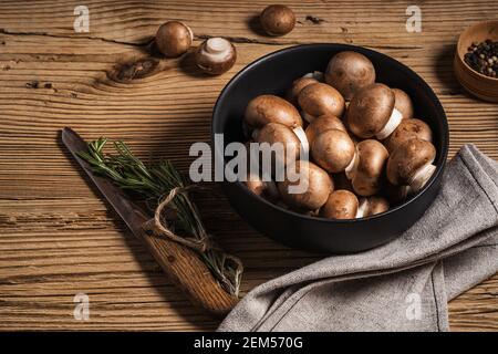 Schüssel mit Baby bella Pilze und Bund von frischem Rosmarin auf rustikalem Holzhintergrund von oben gesehen, pflanzliche Nahrung Stockfoto