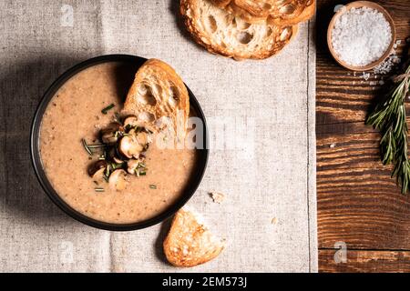 Pilzsahnesuppe serviert mit geröstetem Brot auf Leinen auf rustikalem Holztisch, Blick von oben Stockfoto