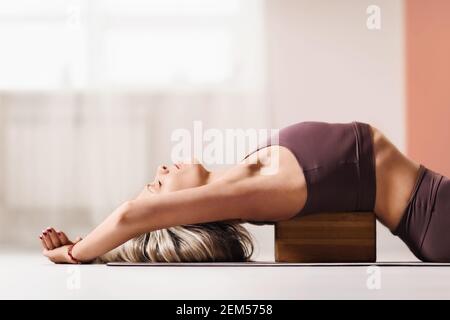 Schöne blonde Frau in Sportswear, liegt auf einem Holzblock für Yoga, um die Haltung zu verbessern. Stockfoto