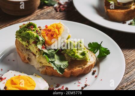 Pochiertes Ei und Avocado mit frischem Koriander auf geröstetem Brot in Scheiben. Toasts auf weißen Esstellern, Brunch für zwei Personen, rustikaler Holzhintergrund Stockfoto