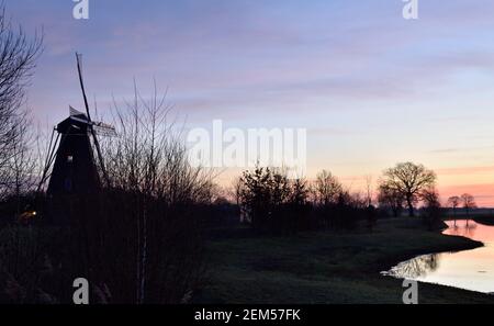 Sonnenaufgang über dem stillen Wasser eines Kanals und Eine typische holländische Windmühle in der Nähe von Vinkel in den Niederlanden Stockfoto