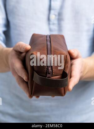 Herren braune Leder persönliche Kosmetiktasche oder Tasche für Toilettenartikel-Zubehör in einem Herrenhänden in blauem Hemd. Stil, Retro, Mode, Vintage und Eleganz. Stockfoto