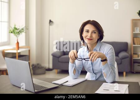 Glückliche Geschäftsfrau, die mit einem Laptop am Schreibtisch sitzt, lächelt und die Kamera anschaut Stockfoto