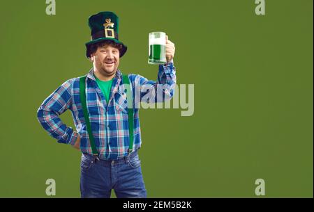 Positiver Mann im karierten Hemd und traditionellen Hut stehend und Hält einen Becher mit grünem Bier Stockfoto