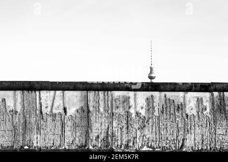 Berliner Mauer original verwitterter Abschnitt beschädigt mit freiliegenden Eisenstangen teilweise den Fernsehturm (Berliner Fernsehturm) weit im Horizont bedeckend. Schwarz und Stockfoto