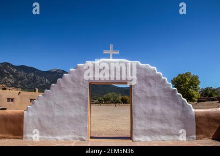 Weiß bemalter Schlamm adobe Tor mit Kreuz auf oben suchen Aus einem Missionshof auf Berge und Bäume und Ein Bewohner des Pueblo mit einem Eimer Witz Stockfoto