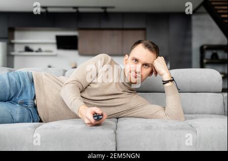 Aufgeregt junger Erwachsener liegt auf einem grauen Sofa mit einer Fernbedienung in der Hand, kann nichts Interessantes im fernsehen finden, auf eine Selbstisolation, zu Hause, nach dem Arbeitstag, leiden an Müßiggang, wurde gefeuert Stockfoto