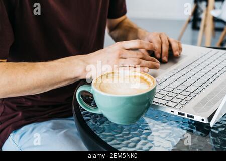 Freiberufliches Lifestyle-Konzept Close up junger Mann Hände auf dem Tisch mit Laptop, Cappuccino und Croissant im Café. Stockfoto