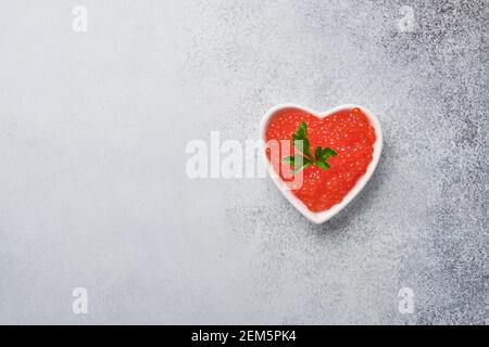 Roter Kaviar in einer herzförmigen Schüssel serviert mit Zitronen- und Eiswürfeln auf einem grauen Tisch. Leerzeichen für Text. Stockfoto