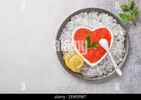 Roter Kaviar in einer herzförmigen Schüssel serviert mit Zitronen- und Eiswürfeln auf einem grauen Tisch. Leerzeichen für Text. Stockfoto