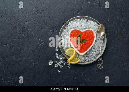 Roter Kaviar in herzförmiger Schüssel serviert mit Zitronen- und Eiswürfeln auf schwarzem Hintergrundtisch. Leerzeichen für Text. Stockfoto