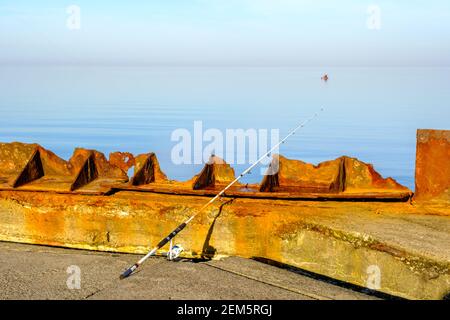 Eine weiße Angelrute hängt über einer Brüstung und der Das Meer ist sehr ruhig Stockfoto