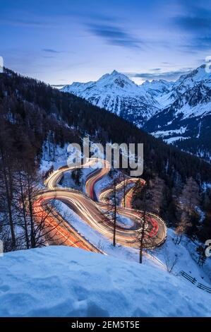 Autos leichte Wege auf den kurvenreichen Kurven der Malojapassstraße im Winter bei Sonnenuntergang, Bregaglia Tal, Kanton Graubünden, Engadin, Schweiz. Stockfoto