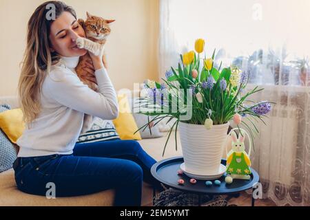 Osterfest zu Hause. Glückliche Frau umarmt Katze entspannend auf Couch. Frühlingsblumen in Topf mit Eiern und Hase auf Couchtisch dekoriert. Stockfoto