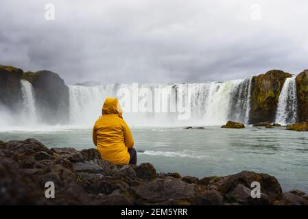 Tourist in einer gelben Jacke, entspannend am Wasserfall Godafoss in Island Stockfoto