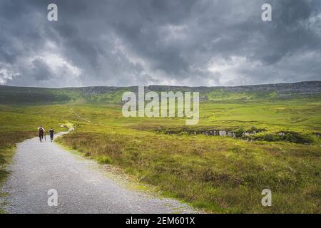 Menschen wandern auf einem Pfad zwischen grünen Hügeln und Moor, der zum Cuilcagh Mountain mit stürmischem, dramatischem Himmel im Hintergrund führt, Nordirland Stockfoto