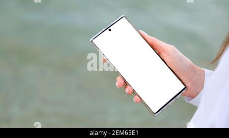 Weißes Mockup Smartphone in weiblichen Händen. Frau verwenden Handy, um im Internet zu suchen, App außerhalb. Nahaufnahme weibliche Hände mit Smartphone auf Meerwasser Stockfoto
