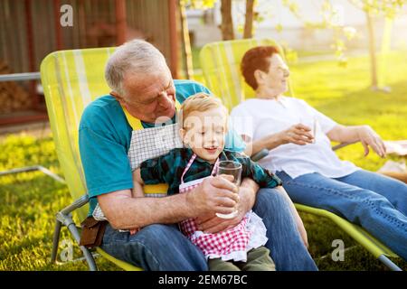 Älterer Mann, der draußen auf einem faulen Stuhl sitzt und seinen herrlich lächelnden Enkel hält und ihm Wasser anbietet. Stockfoto