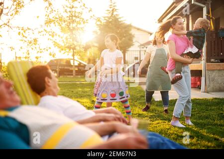 Fröhliche Kinder spielen, während Mutter und Großeltern stolz vor dem Dorfhaus zuschauen. Stockfoto