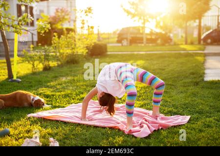 Junge flexible Mädchen führt eine Yoga-Brücke Pose auf einer Decke auf dem Gras, während ihr Hund beobachtet sie. Stockfoto