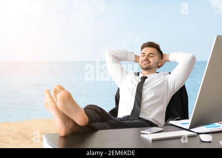 Schöner junger Geschäftsmann, der sich am Arbeitsplatz am Strand entspannt Stockfoto