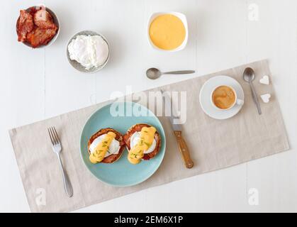 Eier Benedikt, englische Muffins, gegrillter Schinken, pochierte Eier, Sauce Hollandaise, Schnittlauch, Zitrone, Tasse Kaffee. Weißer Holztisch mit Frühstück. Lo Stockfoto