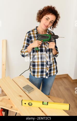 Kaukasische Frau mit lockigen Haaren hält einen Bohrer in den Händen, bereit, um zu Hause Verbesserungen zu machen. Sie steht vor einer Werkbank. Sie trägt einen Plai Stockfoto