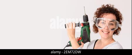 Kaukasische Frau mit lockigen Haaren hält einen Bohrer in den Händen, bereit, um zu Hause Verbesserungen zu machen. Sie trägt ein graues Hemd. Weißer Hintergrund. Tageslicht. Stockfoto