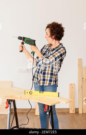 Kaukasische Frau mit lockigen Haaren hält einen Bohrer in den Händen, bereit, um zu Hause Verbesserungen zu machen. Sie steht vor einer Werkbank. Sie trägt einen Plai Stockfoto