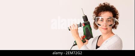 Kaukasische Frau mit lockigen Haaren hält einen Bohrer in den Händen, bereit, um zu Hause Verbesserungen zu machen. Sie trägt ein graues Hemd. Weißer Hintergrund. Tageslicht. Stockfoto