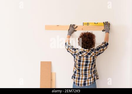 Unkenntlich Frau verwendet eine Ebene, um genau eine Holzdiele auf einer weißen Wand in einer Wohnung mit natürlichem Tageslicht zu platzieren. Stockfoto