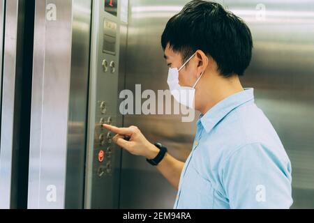 Junge asiatische Geschäftsmann trägt Schutzmaske als Vorsichtsmaßnahme gegen Coronavirus steht im Aufzug und drückt den Boden-Knopf, um nach oben zu gehen Stockfoto