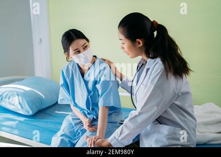 Junge Ärztin mit Stethoskop, das die Hand hält und mit der sie spricht Asiatischer Patient sitzt im Krankenhaus auf dem Bett und trägt ein Sicherheitsgesicht Maske Stockfoto
