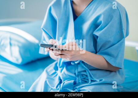 Nahaufnahme der Hände von jungen weiblichen Patienten mit Smartphone zu Text und Surfen im Internet, während Sie auf dem Bett im Krankenhauszimmer sitzen Tragen Peeling Uniform Stockfoto