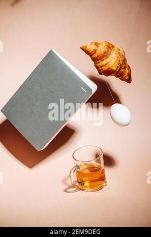 Teetasse auf pfirsichfarbener Oberfläche. Croissant, Ei und Notebook schweben in der Luft Stockfoto