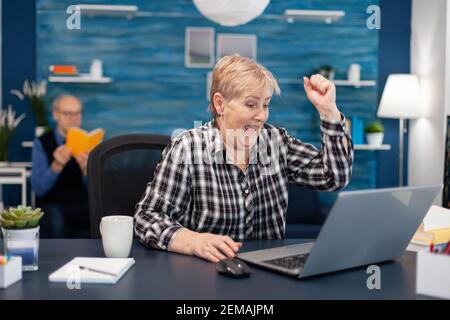 Aufgeregt reife Frau feiert Online-Kauf fröhlich ältere Frau, während mit Laptop-Computer im Wohnzimmer, während der Mann liest ein Buch auf dem Sofa sitzen. Stockfoto