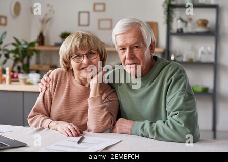 Porträt eines älteren Ehepaars, das die Kamera umarmt und lächelt Sitzen am Tisch mit Laptop und Dokumente zu Hause Stockfoto