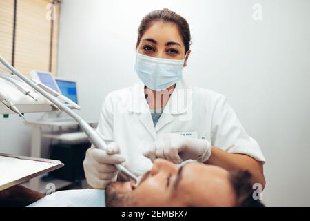 Zahnärztin trägt Gesichtsmaske Blick auf Kamera während der Behandlung eines Patienten. Doctor doing zahnärztliche Behandlung auf die Zähne des Mannes in den Zahnarztstuhl. Stockfoto