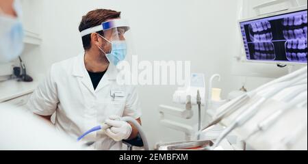 Arzt trägt Schutzschild und Gesichtsmaske Blick auf Röntgenbild des Patienten während der Behandlung. Stockfoto