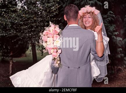 Hochzeit im Jahr 1989 von jungen Modell freigelassene Paar posiert im Freien Für Fotografien in 80s Mode Braut im Hochzeitskleid mit Blumenstrauß gibt Daumen nach oben getragen vom Bräutigam Rückansicht Grauer Morgenanzug und das Tragen einer lächelnden glücklichen Braut historischen 1980s wie wir in Essex England archivierten Stockfoto