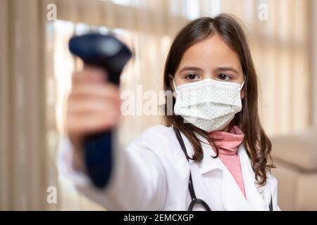 Kleines Mädchen mit Arztuniform und medizinischer Maske mit einem Infrarot-Thermometer. Covid-19 Virenschutzkonzept. Stockfoto