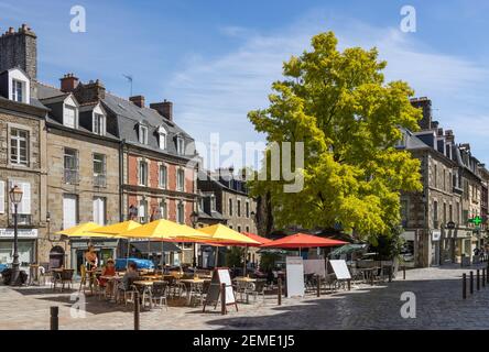 Menschen, die sich in einem Open-Air-Café in Fougeres, Frankreich, entspannen und unter dem Schatten der farbigen Sonnenschirme, umgeben von Gebäuden, den strahlenden Sommersonnen genießen Stockfoto