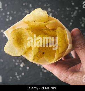 Hintergrund für das Essen. Salzkartoffeln Chips. Hand hält einen Kegel gefüllt von goldenen frittierten Chips . Schnell salzige Lebensmittel für Snack oder Party. Leckeres Essen für den Appetit Stockfoto