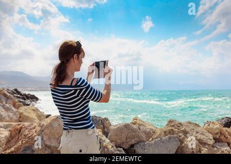 Eine attraktive weibliche Reisende nutzt ihr Handy, um eine schöne Meereslandschaft zu fotografieren. Sonniger Sommertag am Mittelmeer. Speicherplatz kopieren. Stockfoto