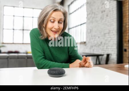 Ältere ältere moderne Frau verwendet Sprachbefehle für drahtlose intelligente Lautsprechersteuerung. Smart Home und Technologiekonzept Stockfoto