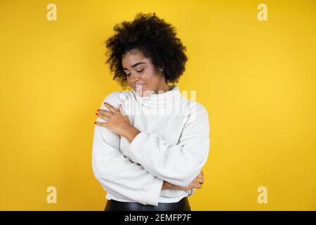 afroamerikanische Frau trägt lässigen Pullover über gelben Hintergrund umarmt sich glücklich und positiv, lächelnd zuversichtlich Stockfoto