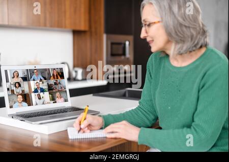 Eine ältere, grauhaarige Dame sitzt zu Hause am Küchentischtisch und studiert online auf einem Laptop, schaut sich ein Webinar mit einer Gruppe an, nimmt an der Online-Konferenz Teil und macht sich Notizen. Videoanruf Stockfoto
