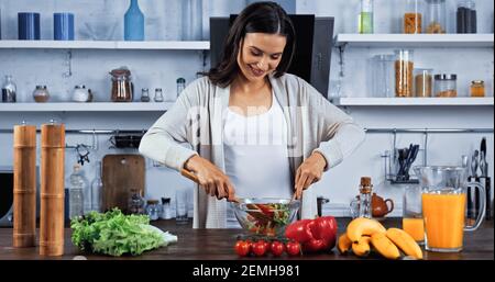 Lächelnde Frau, die Salat in der Nähe frischer Zutaten mischt Stockfoto