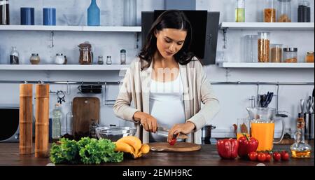 Schwangere Frau schneiden Kirschtomate in der Nähe von frischen Zutaten in der Küche Tabelle Stockfoto