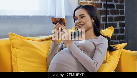 Lächelnde schwangere Frau hält Cupcake auf dem Sofa Stockfoto